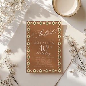 May include: A brown invitation card with the word "Salud!" in elegant script. The card is for Natalie's 40th birthday, with event details and a floral border. White flowers and a candle are in the background.