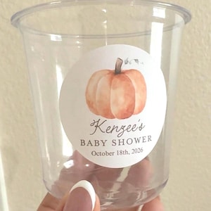 May include: A clear plastic cup featuring a circular white sticker with a watercolor orange pumpkin illustration. The text reads Kenzee's BABY SHOWER October 18th, 2026. The cup has a wide rim and a tapered base, designed for holding beverages or treats.