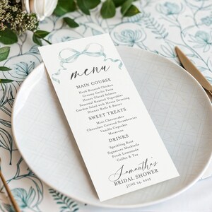 May include: A menu card for Samantha's bridal shower, featuring a watercolor bow design. The menu lists main courses, sweet treats, and drinks. The card is on a white plate with gold cutlery, set on a floral patterned tablecloth.