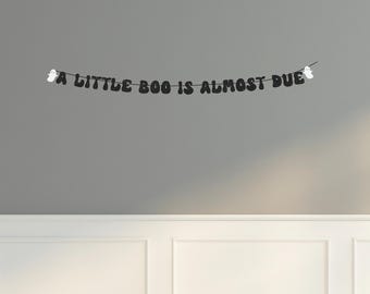 May include: A black banner with the text "A LITTLE BOO IS ALMOST DUE" hangs against a gray wall. The banner has two white ghost decorations, one at each end. The banner is a Halloween-themed decoration.
