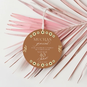 May include: A round, brown thank you tag with white and green floral accents. The tag reads "MUCHAS gracias POR CELEBRAR CONMIGO MI CUMPLEAÑOS! NATALIE 40 Salud! JUNE 12, 2024".