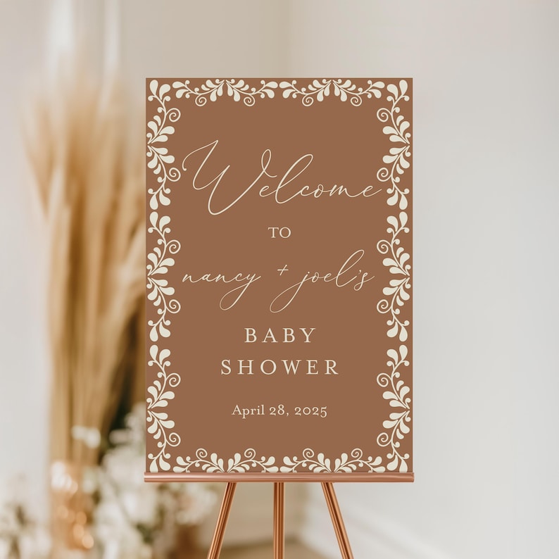 May include: A brown welcome sign with a white floral border. The sign reads "Welcome to Nancy + Joel's Baby Shower April 28, 2025".