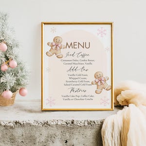 Op de afbeelding: Een ingelijst menu met een gouden rand, met een peperkoekmannetje-illustratie en de tekst "MENU". Het menu bevat ijskoffie, add-ons en gebak. Links staat een kleine kerstboom met roze ornamenten.