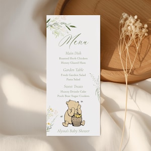 Può includere: Un biglietto del menu bianco con un design floreale e la parola "Menu" in corsivo. Il menu elenca piatti come pollo alle erbe arrosto e torta al miele. Un'illustrazione di Winnie the Pooh è in basso, con il testo "Alyssa's Baby Shower."