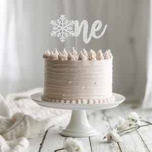 May include: A white cake with pink frosting and a snowflake and "one" cake topper. The cake is on a white cake stand and there are white flowers on a white wooden table.