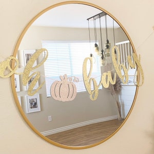 May include: A gold glitter banner with the words "oh baby" and a pink glitter pumpkin in the middle. The banner is hanging in front of a mirror.