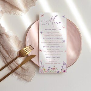 Könnte beinhalten: Eine Menükarte mit floralem und Schmetterlingsdesign, mit Menüpunkten wie Vorspeisen, Brunch-Auswahl, süßen Genüssen und Erfrischungen. Die Speisekarte liegt auf einem rosa Teller mit goldenem Besteck und einem beigen Tuch.