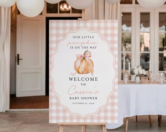 May include: A rectangular baby shower welcome sign featuring a light pink and white gingham background. The center displays a watercolor pumpkin with a pink bow, and text reading: Our Little Pumpkin Is On The Way, Welcome to Cassie's Baby Shower, October 27, 2026.