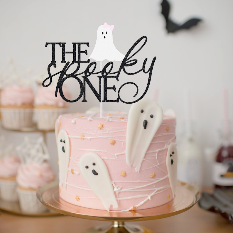 May include: A pink and white cake decorated with ghost-shaped fondant and a black glitter topper that reads "The Spooky One".