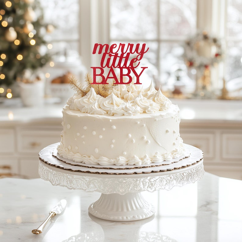 May include: White cake with white frosting and red glitter 'merry little baby' topper on a white cake stand.