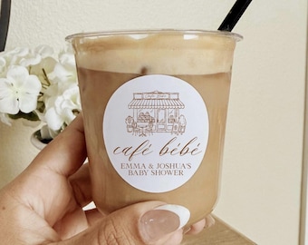 May include: A clear plastic cup filled with iced coffee topped with foam and a black straw. A circular white sticker on the front features a brown line drawing of a cafe storefront with the text café bébé and EMMA & JOSHUA'S BABY SHOWER.