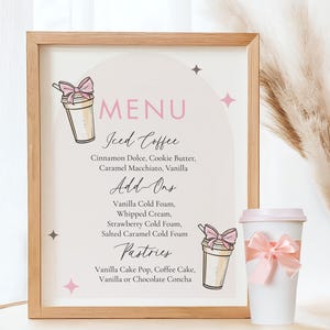 May include: A framed menu board with a light wood frame. The menu features "MENU" in pink, with "Iced Coffee" and "Add-Ons" listed below. A white coffee cup with a pink lid and bow is on the right. The menu lists coffee flavors and pastries.