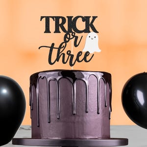May include: A chocolate cake with dark chocolate drips, a black glitter cake topper that reads "Trick or Three", and a white ghost. Two black balloons are on either side of the cake. The cake is on a black cake stand.