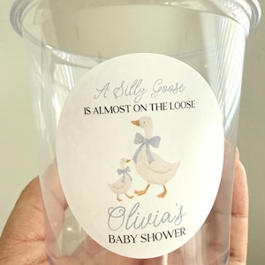 May include: A clear plastic cup featuring a circular sticker with a watercolor illustration of two geese wearing blue bows. The text reads: A Silly Goose IS ALMOST ON THE LOOSE, Olivia's BABY SHOWER. The design is centered on the front of the cup.