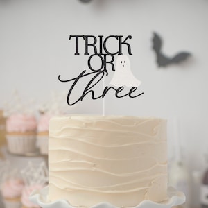 May include: A white cake with black frosting and a black and white "Trick or Three" cake topper with a ghost.