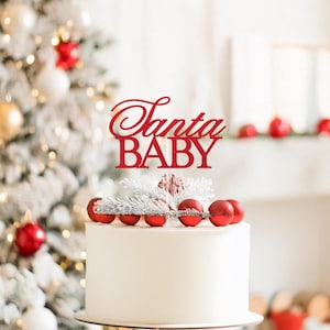 May include: A white cake topped with red Christmas ornaments and a red cake topper that reads "Santa BABY". The cake is decorated with silver and white accents. A Christmas tree is in the background.