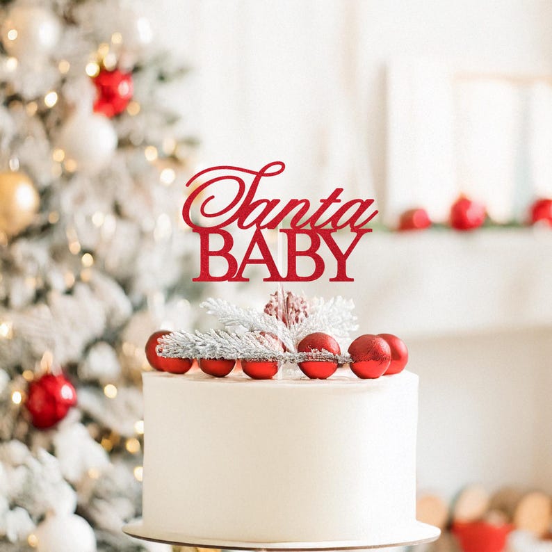 May include: A white cake topped with red Christmas ornaments and a red cake topper that reads "Santa BABY". The cake is decorated with silver and white accents. A Christmas tree is in the background.