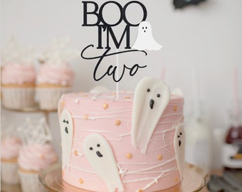 May include: A pink cake decorated with white ghost figurines and a black cake topper that reads "BOO I'M TWO".