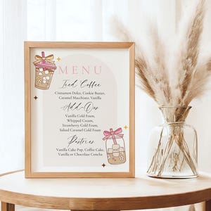 May include: A framed menu board with a light wood frame, displaying a menu for iced coffee and pastries. The menu includes options like Cinnamon Dolce, Vanilla, and Vanilla Cake Pop. The board is on a round wooden table.