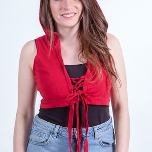 May include: A red hooded crop top with a lace-up front and long sleeves. The top is tied at the waist and worn over a black tank top and blue denim shorts.