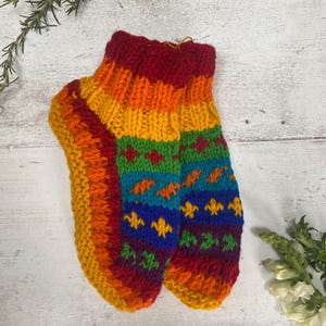 May include: A pair of hand-knitted ankle socks featuring a vibrant rainbow color palette. The socks display horizontal stripes in red, orange, yellow, green, blue, and purple, accented with small, repeating geometric patterns in contrasting yarn colors.