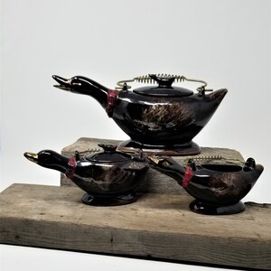 May include: A brown and black ceramic teapot, creamer, and sugar bowl set shaped like ducks. The teapot has a spring-loaded lid and a gold handle.