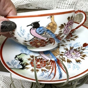 May include: A white porcelain teacup and saucer set with a hand-painted design of birds and flowers. The birds are depicted in shades of blue, pink, and yellow, while the flowers are red, pink, and gold.