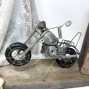 May include: A handcrafted wire sculpture of a motorcycle. The motorcycle features a black tire, silver wire frame, and a detailed design. The sculpture is displayed on a wooden surface, showcasing the intricate craftsmanship of the wire art.