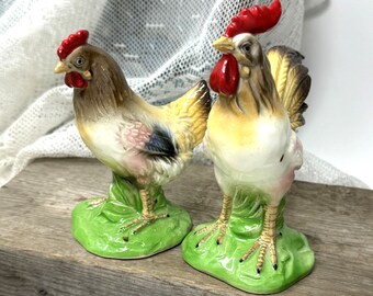 May include: A pair of ceramic rooster and hen figurines. The rooster is brown and white with a red comb and wattle. The hen is brown and white with a pink and blue wing. Both figurines are standing on a green base.
