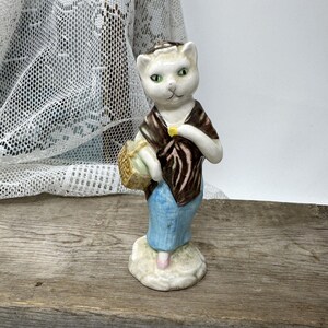 May include: A ceramic figurine of a cat-like figure wearing a shawl and blue pants, holding a basket. The cat has green eyes and a brown and white patterned shawl. The figurine is standing on a small, textured base.