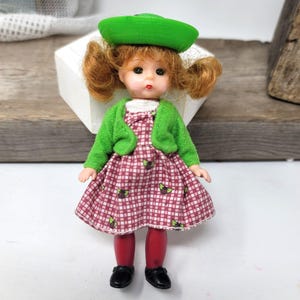 May include: A small doll wearing a green hat, a green cardigan, a red and white plaid dress, and red tights. The doll has red hair and blue eyes.