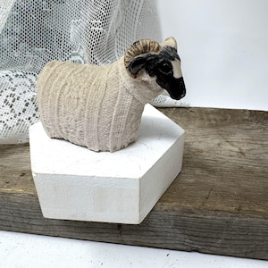 May include: A small figurine of a ram with a black face and curved brown horns. The body of the ram is textured to resemble a cream-colored knitted sweater with vertical ribbing patterns, creating a distinct contrast between the smooth face and wool.