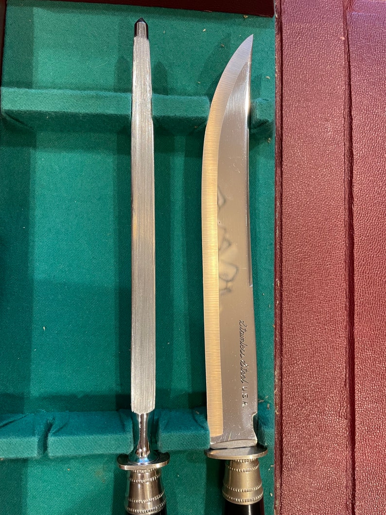 Pu&ograve; includere: Primo piano di due coltelli da cucina argentati con manici neri. Il coltello a sinistra &egrave; un acciaio per affilare e il coltello a destra &egrave; un coltello da intaglio. Il coltello da intaglio ha la scritta "Stainless Steel U.S.A." incisa sulla lama.