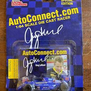 May include: A blue 1:64 scale die-cast race car with a black and white checkered pattern on the side. The car has a Ford logo and the number 51 on the side. The car is on a blue and white card with the text "AutoConnect.com 1998 Special Edition" and a signature from Greg LeMond.