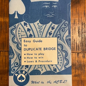Può includere: Un libretto blu e bianco intitolato "Easy Guide to Duplicate Bridge" con un'illustrazione di una carta da gioco di picche nera sulla copertina. Il libretto contiene informazioni su come giocare, come vincere e le leggi e le procedure del gioco. Il logo dell'American Contract Bridge League (ACBL) è presente nell'angolo in basso a sinistra della copertina.