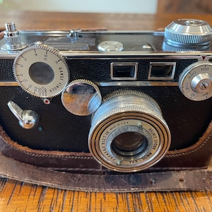Może przedstawiać: Czarno-srebrny aparat fotograficzny 35 mm z skórzanym futerałem. Aparat posiada dalmierz, obiektyw z obiektywem Argus Cintar 7,5-50 mm i pokrętło prędkości migawki. Aparat jest w dobrym stanie i wydaje się być vintage.