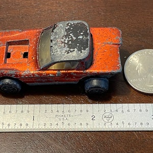 Pode incluir: Carro de brinquedo laranja com teto preto e compartimento do motor visível. O carro está ligeiramente desgastado e tem um acabamento de pintura desgastado. O carro mede aproximadamente 5 cm de comprimento e está sobre uma superfície de madeira ao lado de uma régua e uma moeda de vinte e cinco centavos.