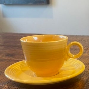 Puede incluir: Un juego de taza y platillo de té de cerámica amarilla. La taza tiene una forma redondeada y un asa simple. El platillo tiene un borde festoneado.