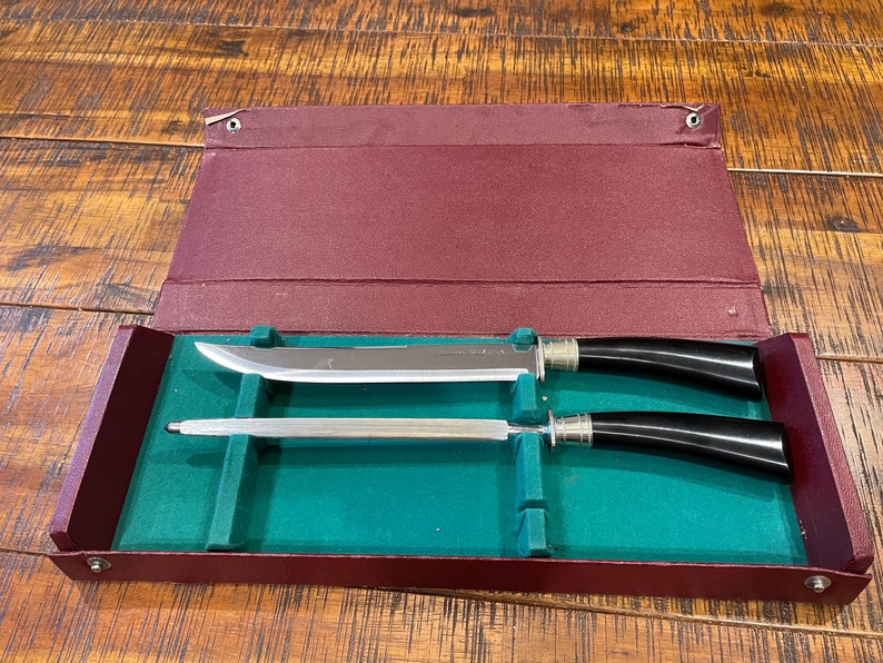 Pu&ograve; includere: Un set di due coltelli d'argento con manici neri in una custodia di velluto bordeaux e verde.