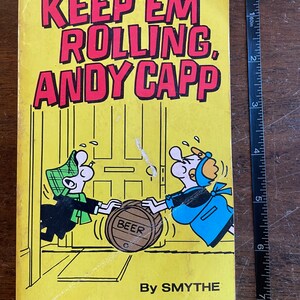 May include: A yellow comic book cover with the title "Keep 'Em Rolling, Andy Capp" in red letters. The cover features a cartoon illustration of two people pushing a barrel labeled "Beer".