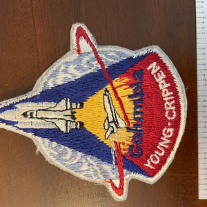 May include: A blue, white and red embroidered patch with the words "Columbia" and "Young-Crippen" in white. The patch depicts a space shuttle launching into space with a yellow sun and a red and white earth in the background.