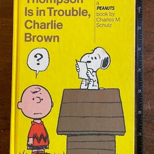 Puede incluir: Una cubierta de libro amarilla con el título "Thompson Is in Trouble, Charlie Brown" y una ilustración de Peanuts de Charlie Brown y Snoopy. El libro es un libro de Peanuts de Charles M. Schulz.