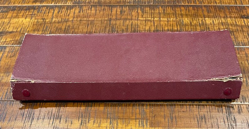 Pu&ograve; includere: Una scatola rettangolare di colore bordeaux con bordi arrotondati. La scatola ha due piccoli bottoni rotondi di colore rosso scuro sul davanti.