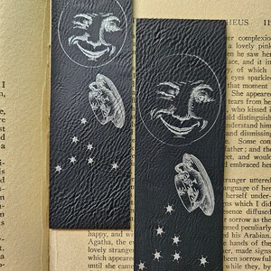 May include: Two black leather bookmarks with a white moon and stars design. The moon is smiling and winking. The bookmarks are decorated with a textured leather surface.