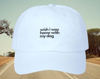 May include: White baseball cap with the text "wish i was home with my dog" in black. The hat has a curved brim and a rounded crown. The background shows a road and a light blue sky.