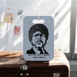 May include: A rectangular, light gray luggage tag featuring a black and white image of a man's face and the text "DID YOU JUST TOUCH RON'S BAG?!" A white cord is attached for securing the tag to luggage. The tag is a travel accessory.