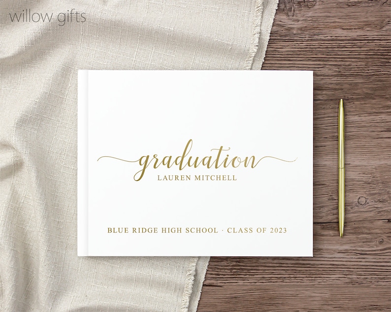 May include: White graduation guest book with gold lettering. The book says "graduation" with the name "Lauren Mitchell" below. The book also says "Blue Ridge High School - Class of 2023".