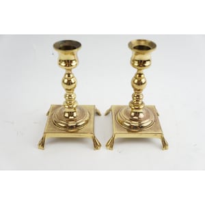 May include: A pair of polished brass candlesticks featuring a tall, slender stem with rounded decorative nodes, a flared top cup, and a square base supported by four small, curved feet. The metal surface has a bright, reflective gold-toned finish.