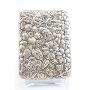 May include: A rectangular silver-toned case with a detailed floral design. The case is covered in embossed flowers, leaves, and buds. The flowers are of various sizes and shapes, creating a textured surface.