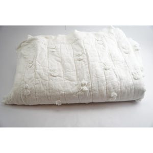 May include: A folded white quilt with a textured surface and vertical stitching. The quilt features small, raised floral-like embellishments scattered across its surface. The overall design is simple and elegant, suitable for a bedroom or living room.